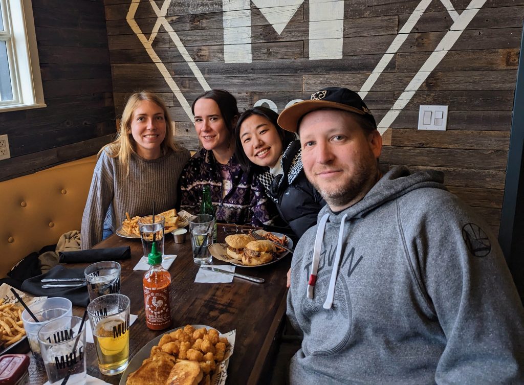 Four people sitting around a table with food and drinks. A large M on the wall is the restaurant logo